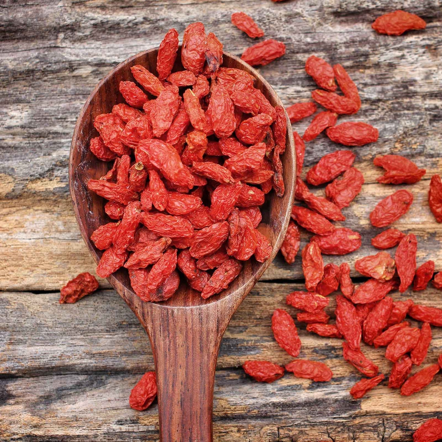 Close-up of goji berries inside a wooden spoon, spilling out on a wooden background.