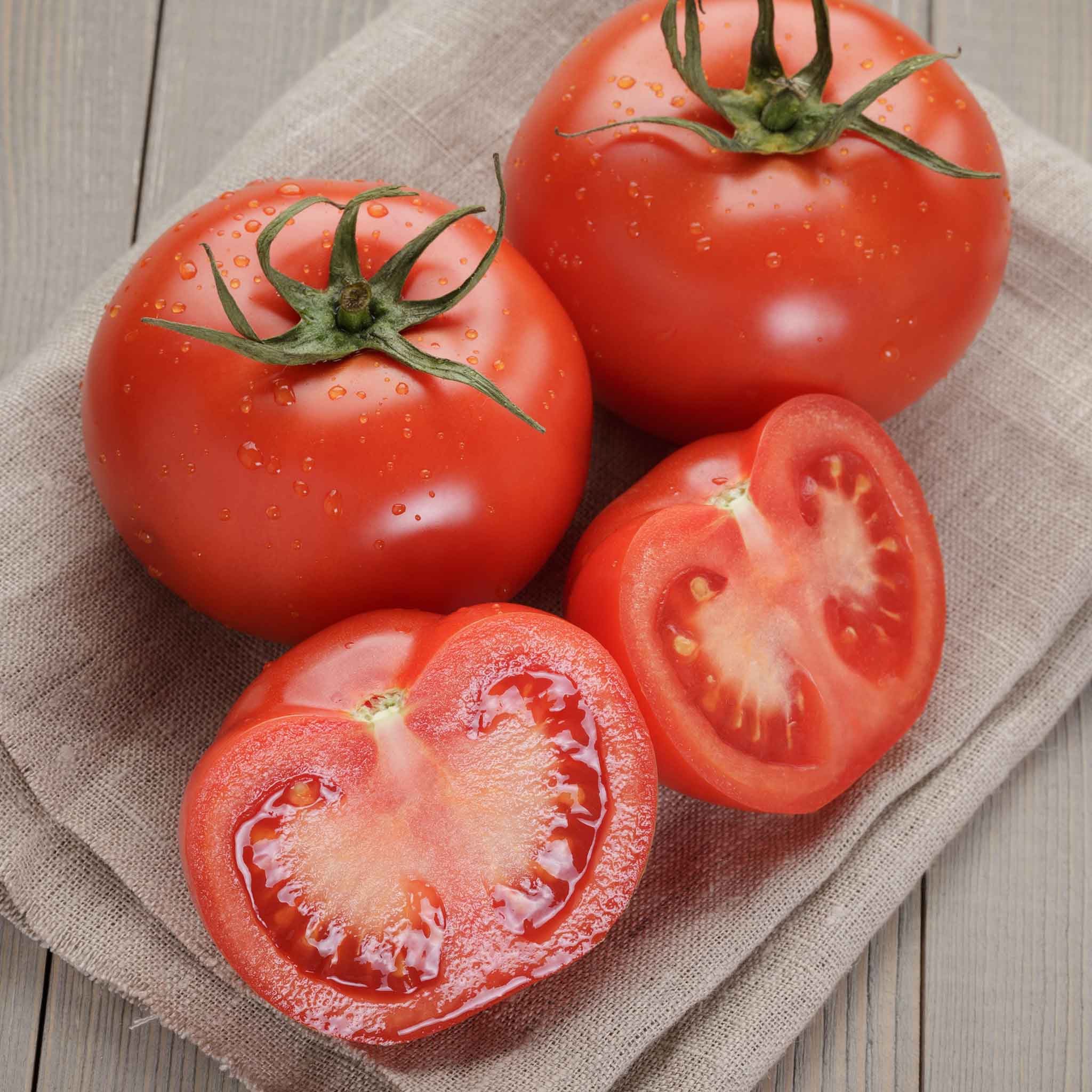 A ripe, red Homestead tomato with a green stem still attached, sitting on a wooden surface with some sliced tomatoes.
