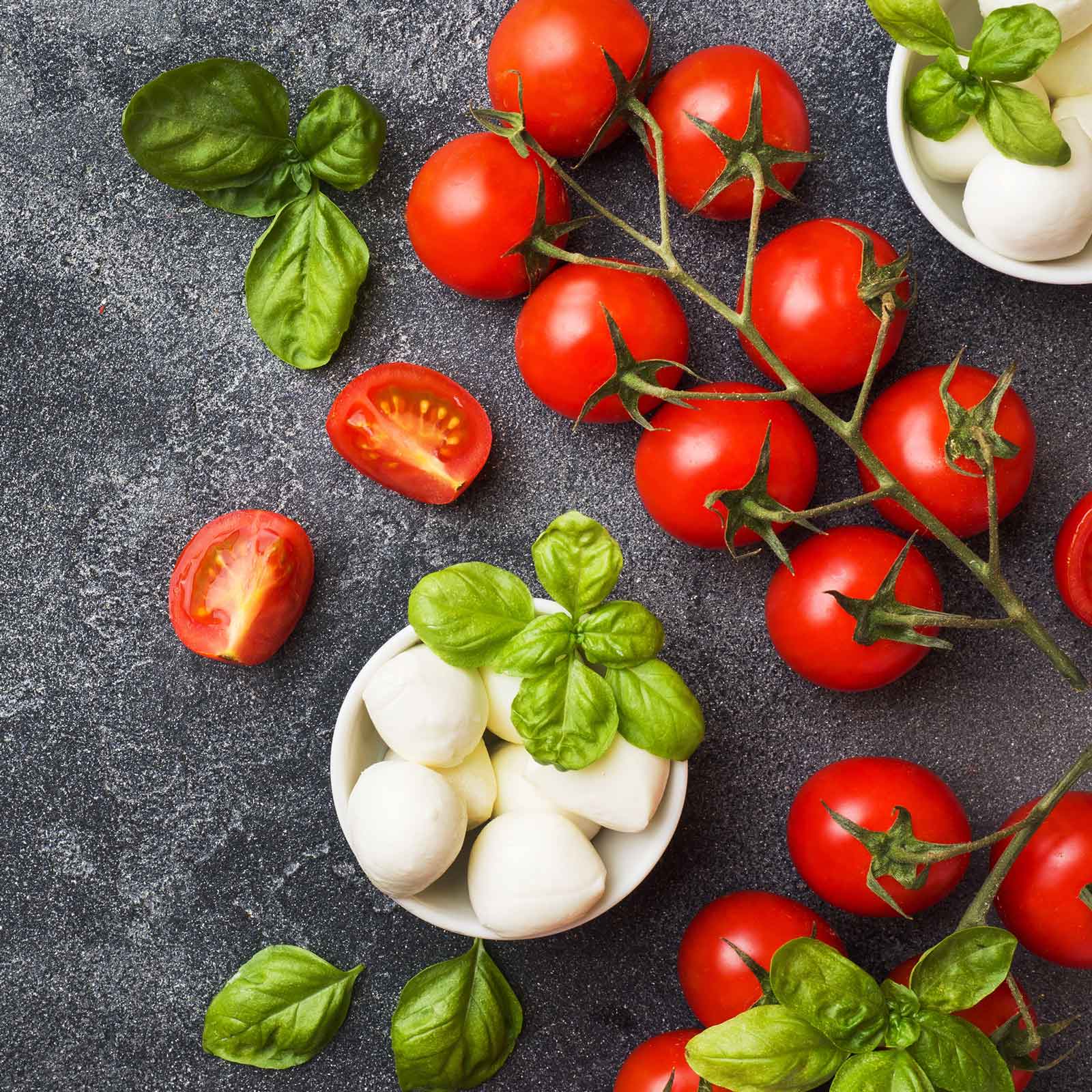 A cluster of small red cherry tomatoes on the vine, with green stems next to a bowl of mozzarella.