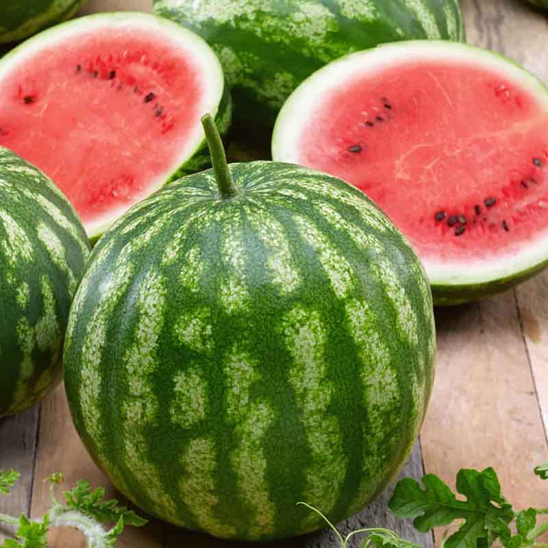 A Cal Sweet watermelon with dark green stripes on a wooden plank with two halves of watermelon in the background.