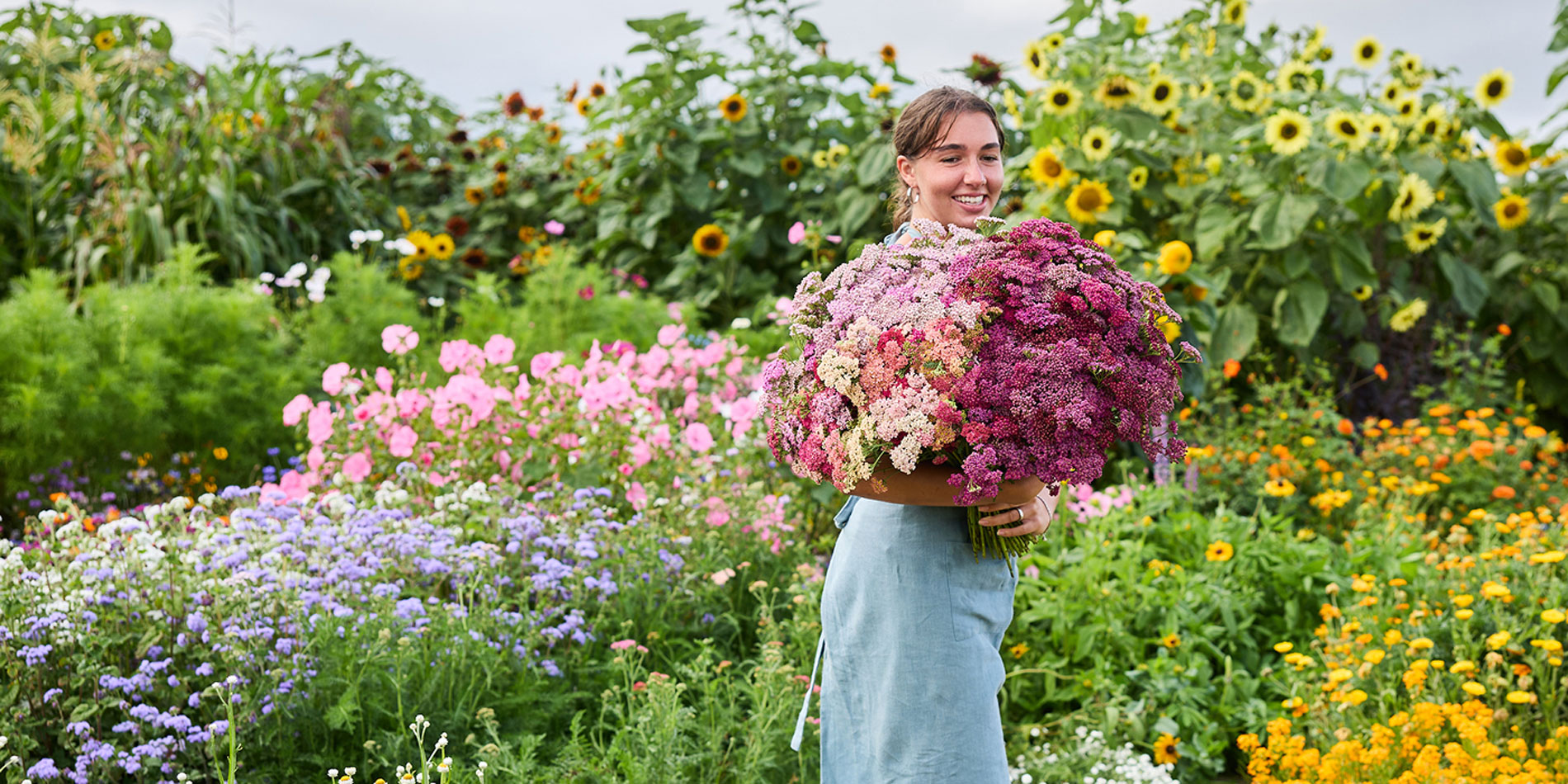 A woman standing in a vibrant flower garden, holding a large, dense bouquet of pink, purple, and cream wildflowers.