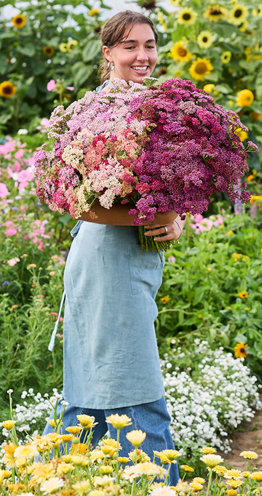 A smiling woman in a blue apron stands in a sunlit flower garden holding a very large, round bouquet of pink, purple, and white wildflowers.