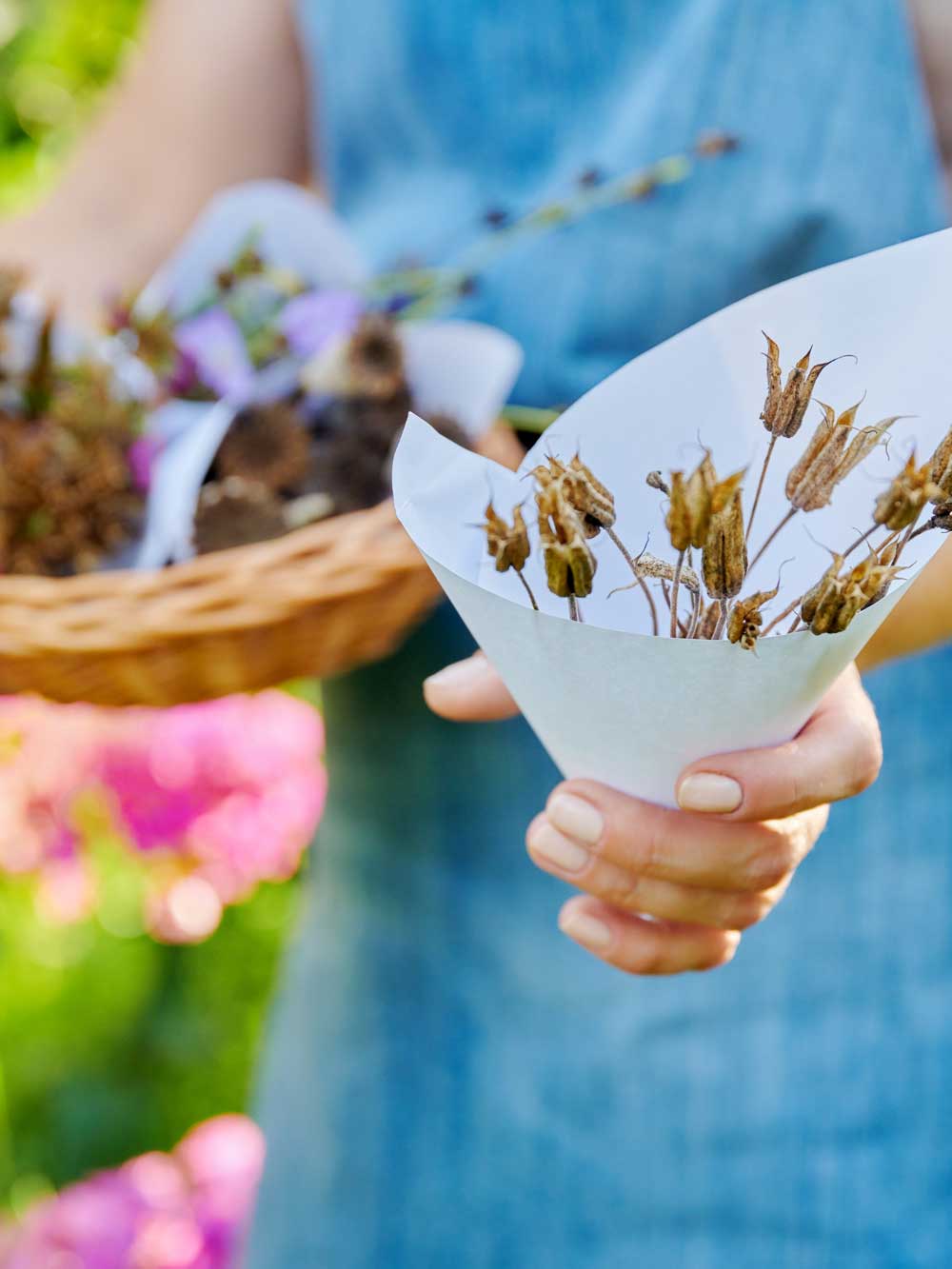 A person in a blue dress holds a cone of dried flowers or seed pods in one hand and a wicker basket in the other, with blurred pink flowers and greenery in the background.