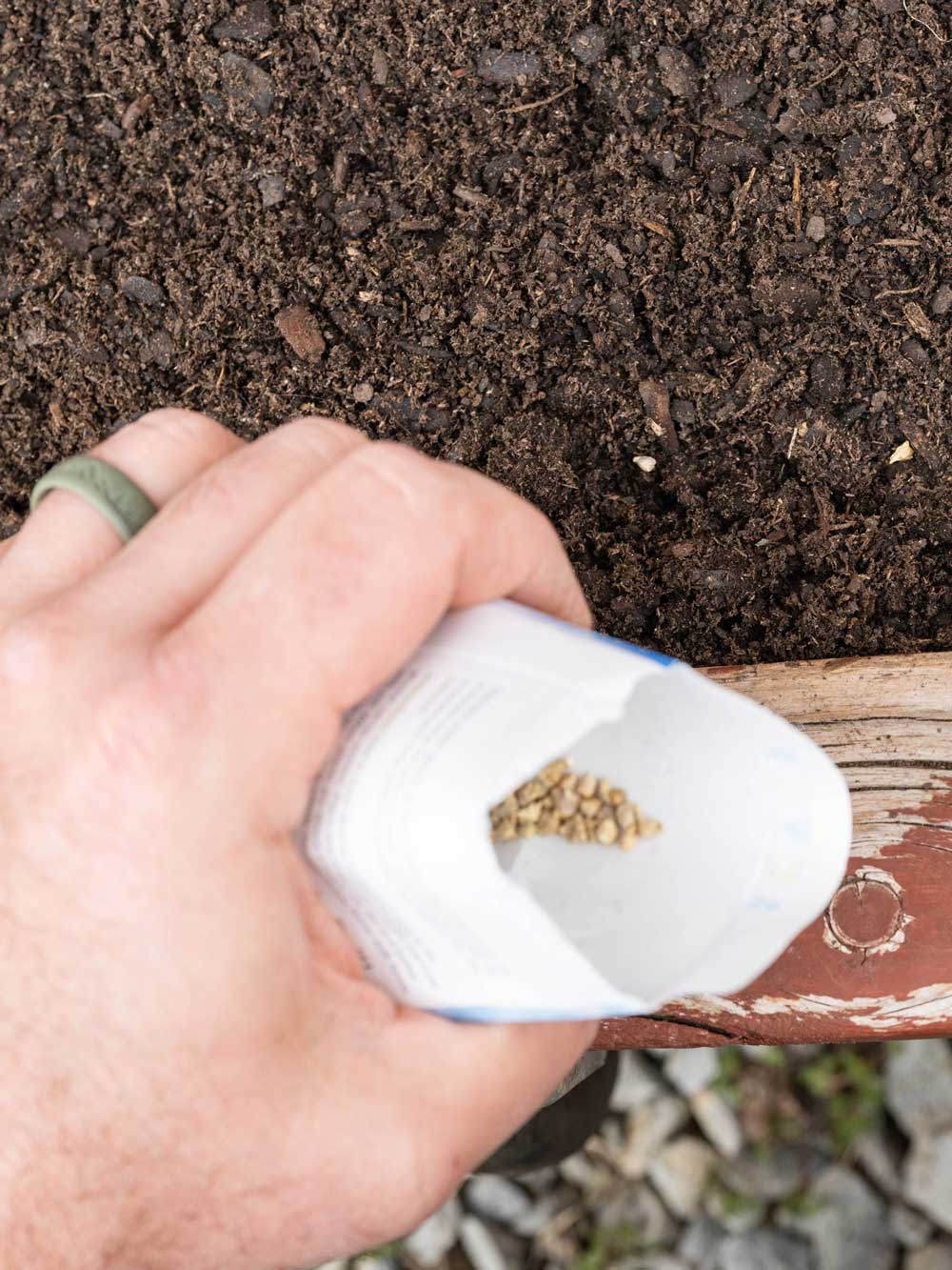 A hand holding an open white paper bag over dark soil, with small tan seeds visible inside the bag.