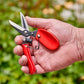 Hand holding a pair of red-handled comfortgel dead headers against a blurred green background