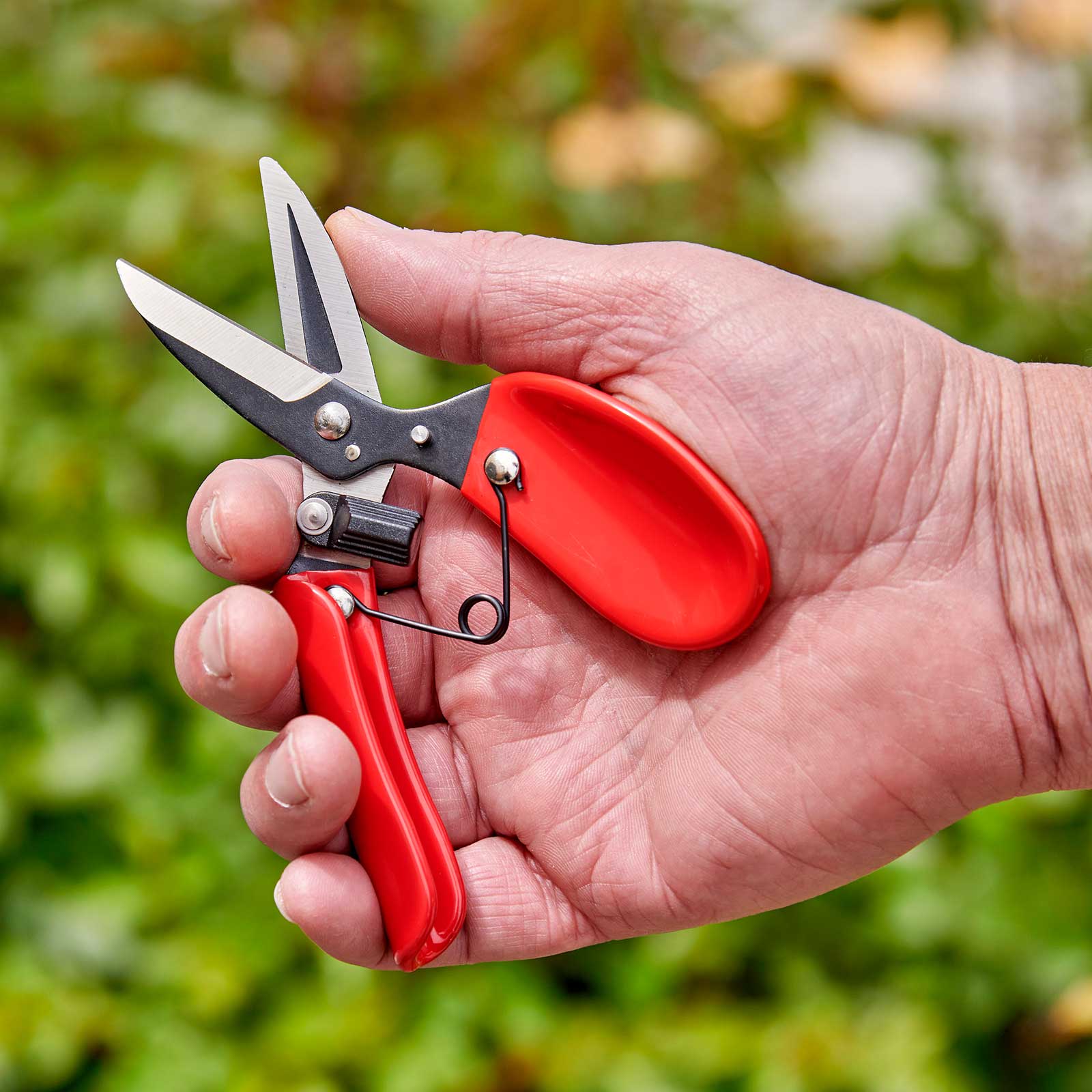 Hand holding a pair of red-handled comfortgel dead headers against a blurred green background