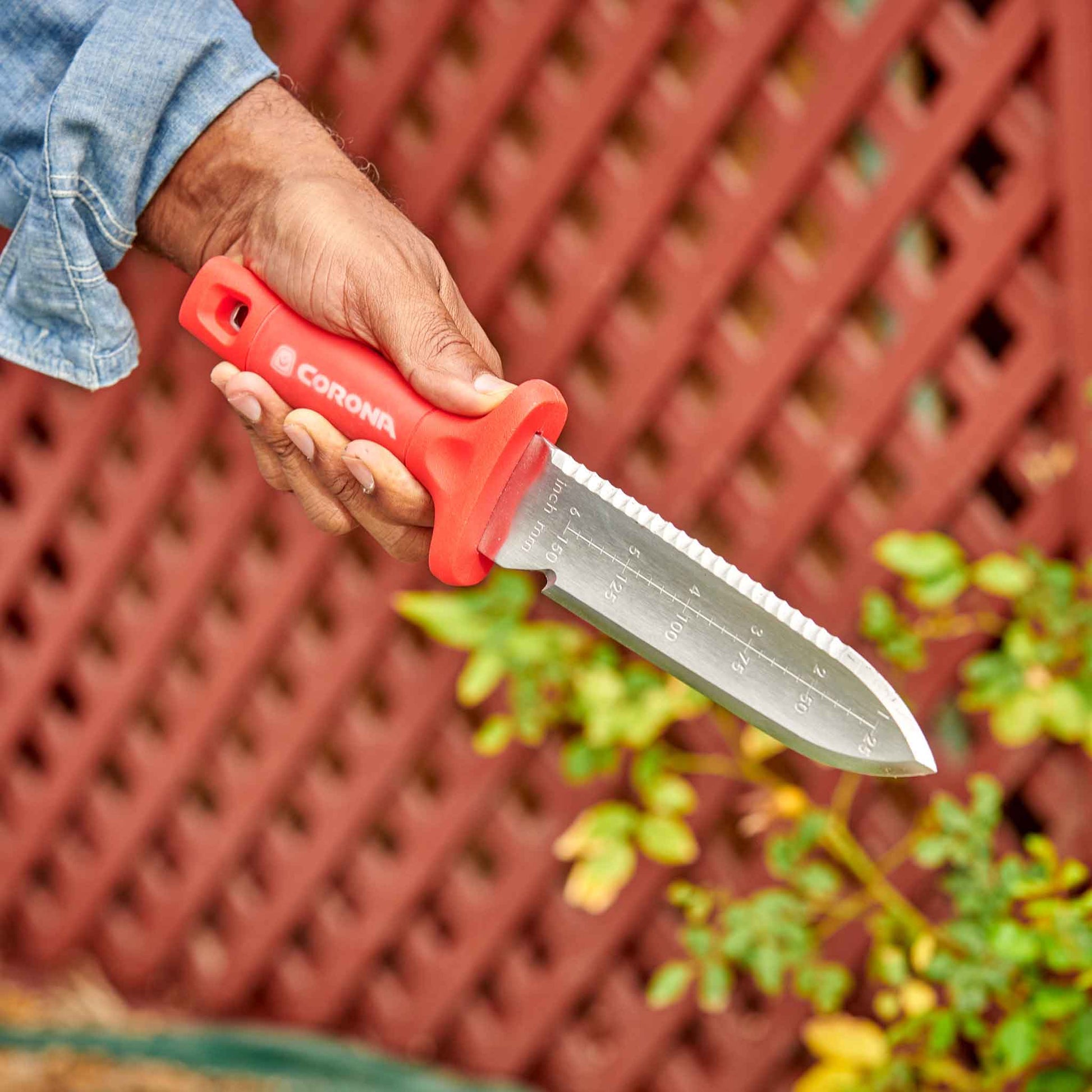 Hand holding a Corona ComfortGel Hori Hori garden knife against a red lattice background