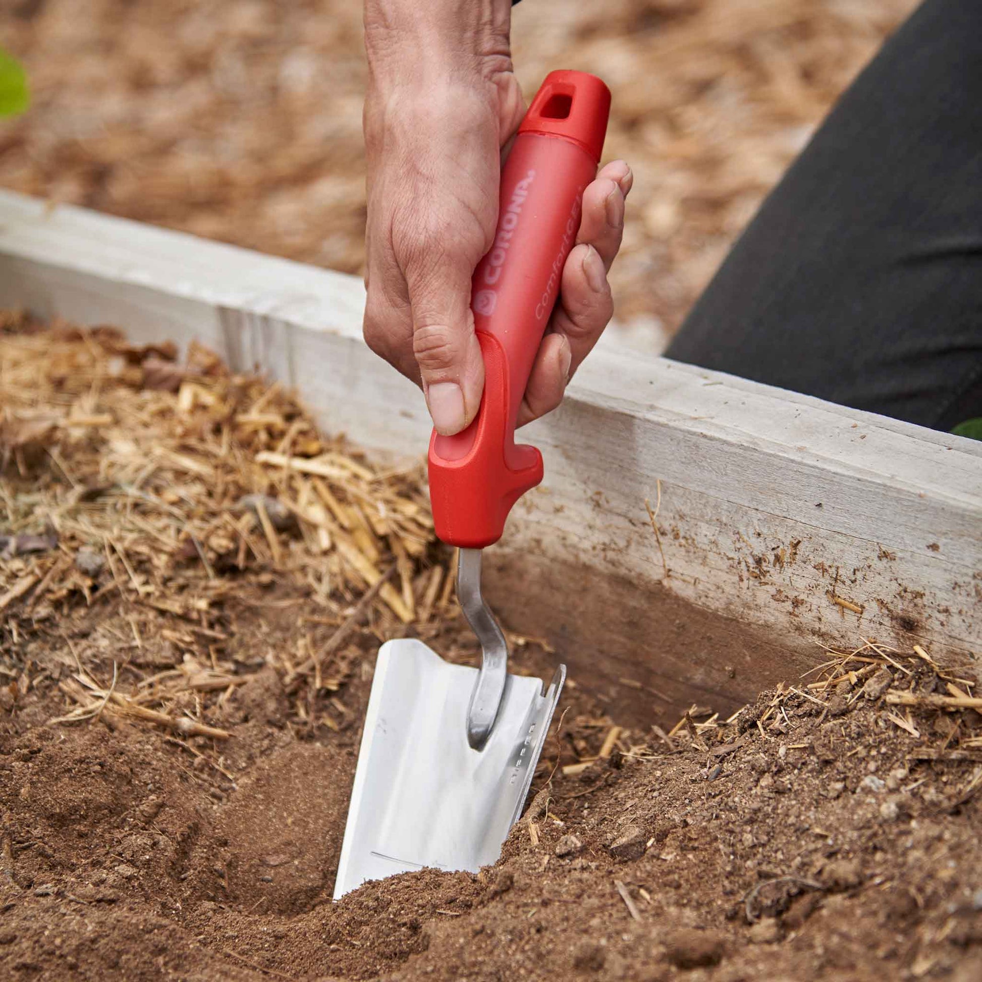 Hand using a red-handled corona comfortgel trow to dig into soil in a raised bed.