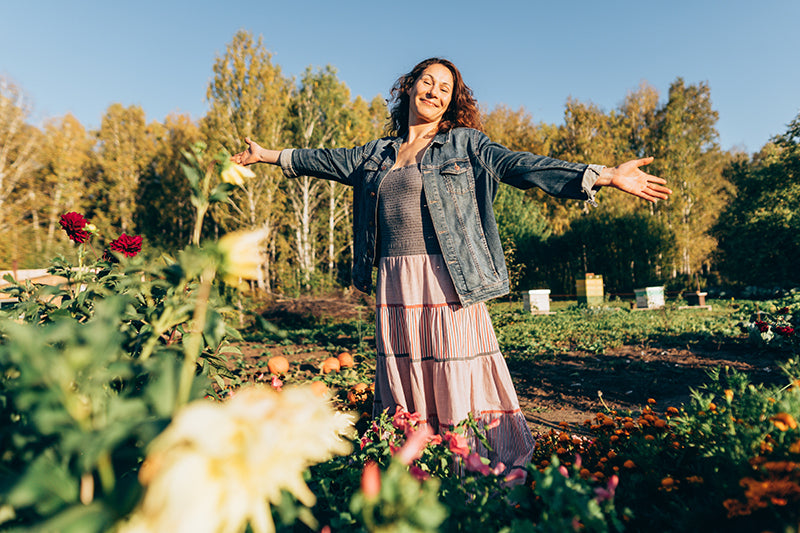 A woman stands in a sunny garden with her arms outstretched, wearing a denim jacket over a gray top and a long pink tiered skirt. Trees and flowers fill the background and foreground.