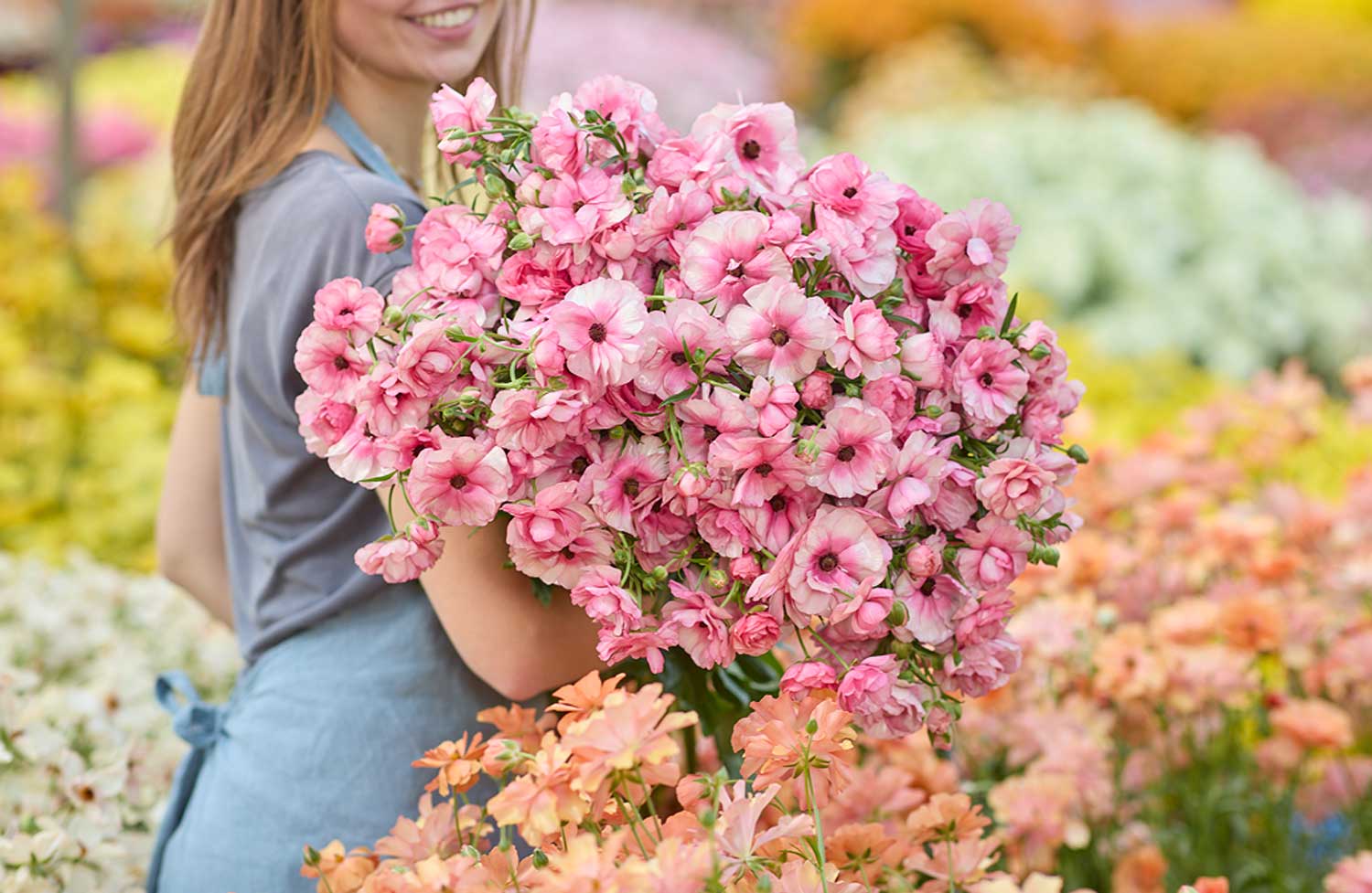 A smiling woman in a garden holds a large bouquet of pink petunia flowers. The background is a soft-focus colorful flower field with yellow, orange, and white blossoms.