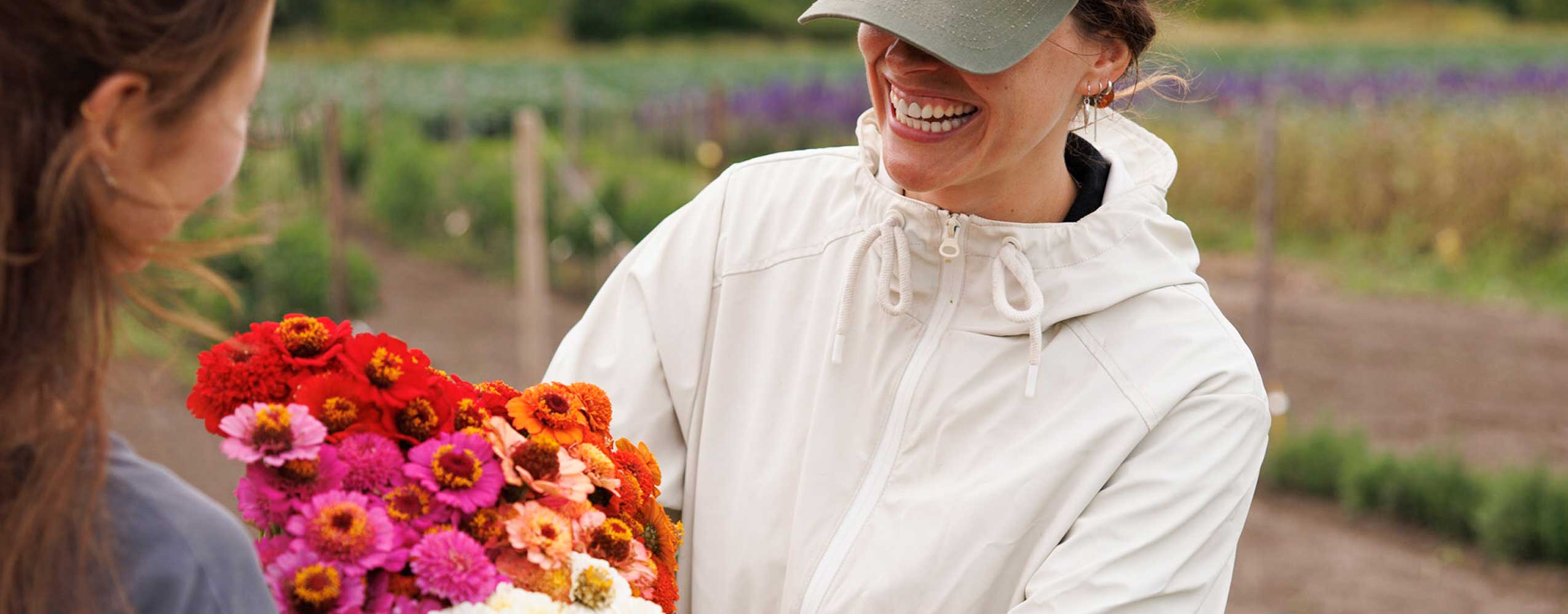 A woman in a white hooded jacket and green cap smiles while holding a large bouquet of colorful flowers, standing outdoors in a garden or field.
