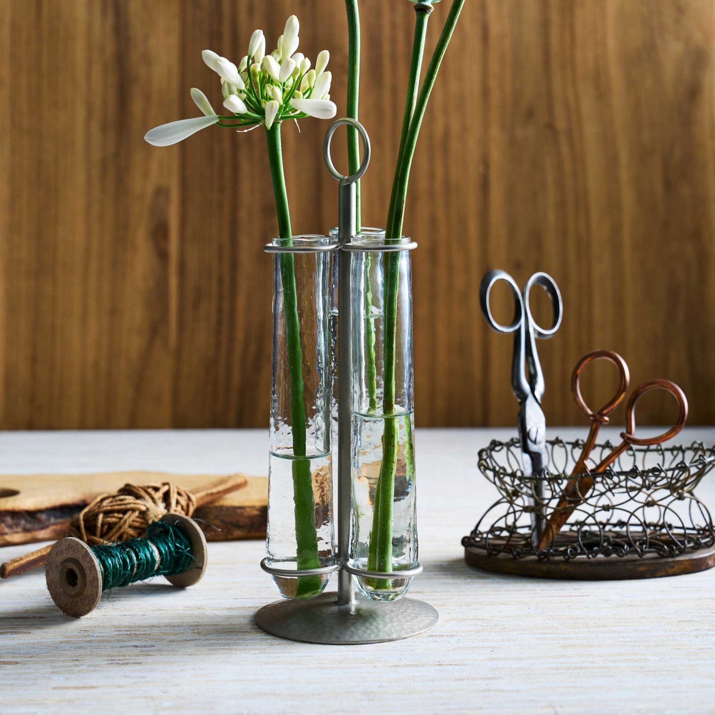 The collective glass bud vase made from two upright test tubes connected by a silver metal frame and base, holding green stems and white flowers, styled on a light wood surface with scissors and spools in the background.