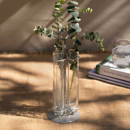 The collective glass bud vase with a silver metal base, holding green eucalyptus stems, set on a textured beige fabric surface with warm natural light and soft shadows.