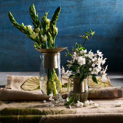The Collective Hammered Glass Arranging Vases with metal flower guards, one holding green asparagus and the other white jasmine blossoms, arranged on a patterned beige cloth against a textured blue background.