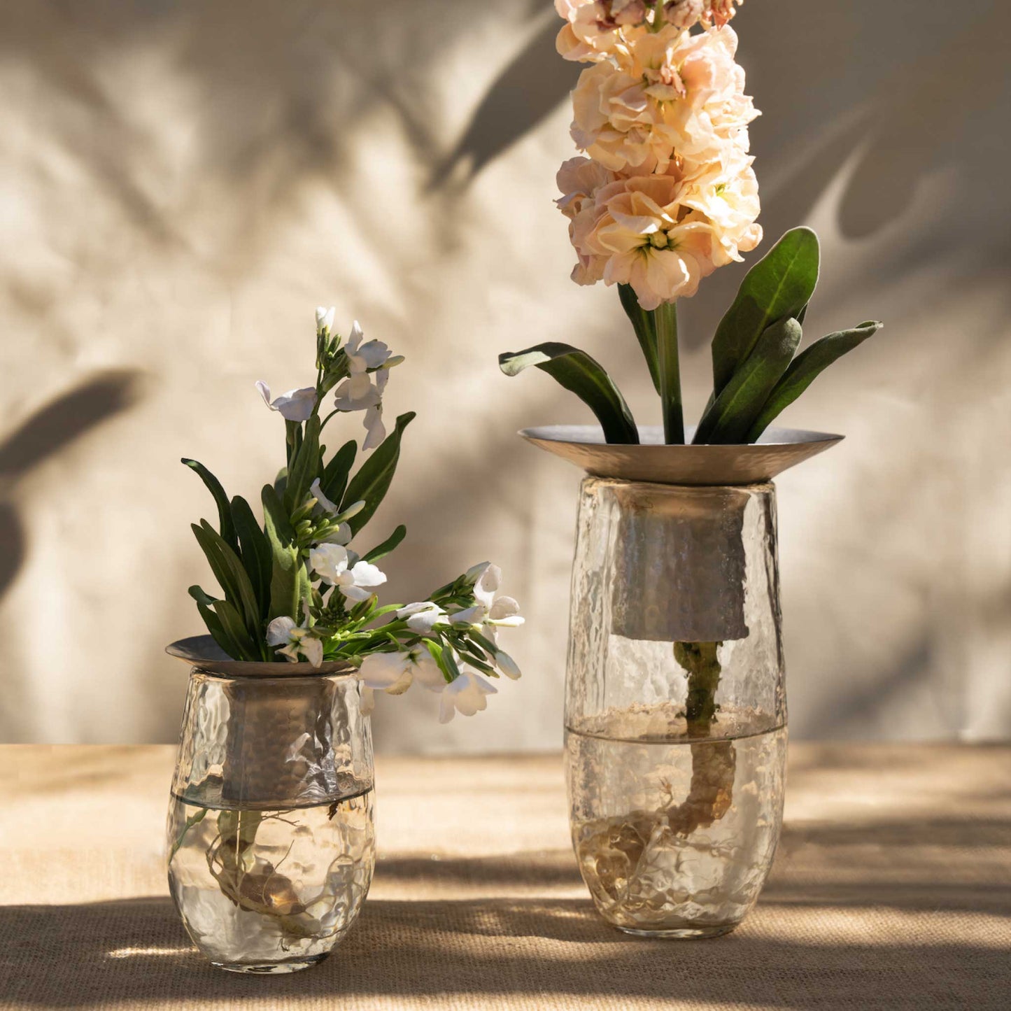 The Collective Hammered Glass Arranging Vases with metal flower holders, filled with dark burgundy artificial flowers, set on a light wood surface against a textured off-white wall.