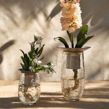 The Collective Hammered Glass Arranging Vases with metal flower holders, filled with dark burgundy artificial flowers, set on a light wood surface against a textured off-white wall.