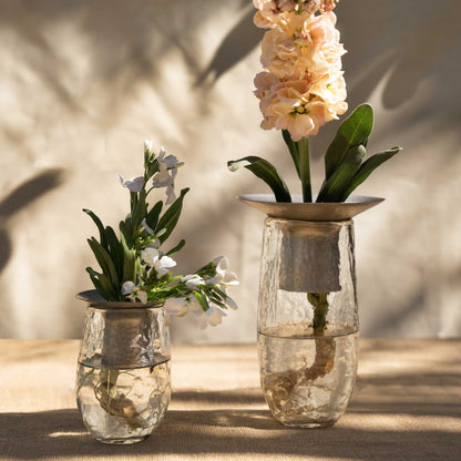 The Collective Hammered Glass Arranging Vases with metal flower holders, filled with dark burgundy artificial flowers, set on a light wood surface against a textured off-white wall.