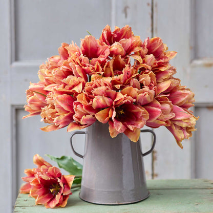Bouquet of pink and orange callahan tulip flowers in a metal vase on a wooden surface with a light gray background.