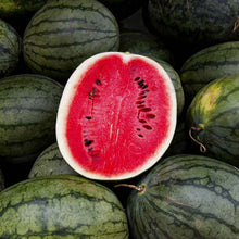 A cross-section of a Black Diamond watermelon shows its red flesh and black seeds on a pile of other Black Diamond watermelon.